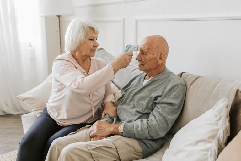 Senior woman checking her partner's temperature on a sofa, promoting health care.